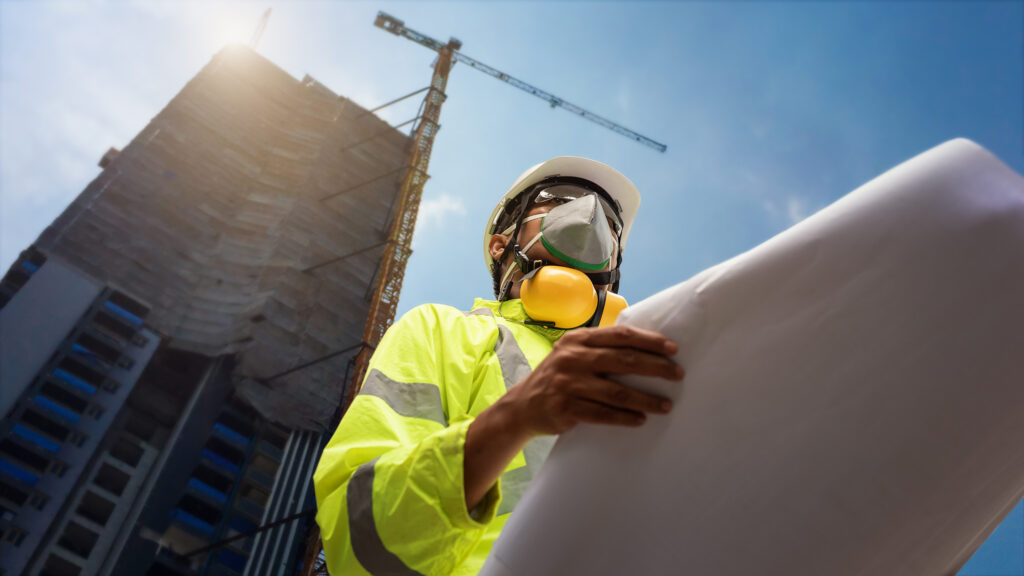 engineer wearing mask holding up building plans in front of skyscraper