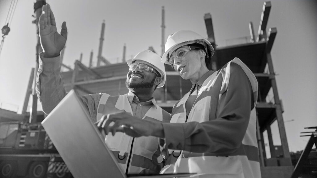 Two Specialists Inspect Commercial, Industrial Building Construction Site. Real Estate Project with Civil Engineer, Investor Use Laptop. In the Background Crane, Skyscraper Concrete Formwork Frames