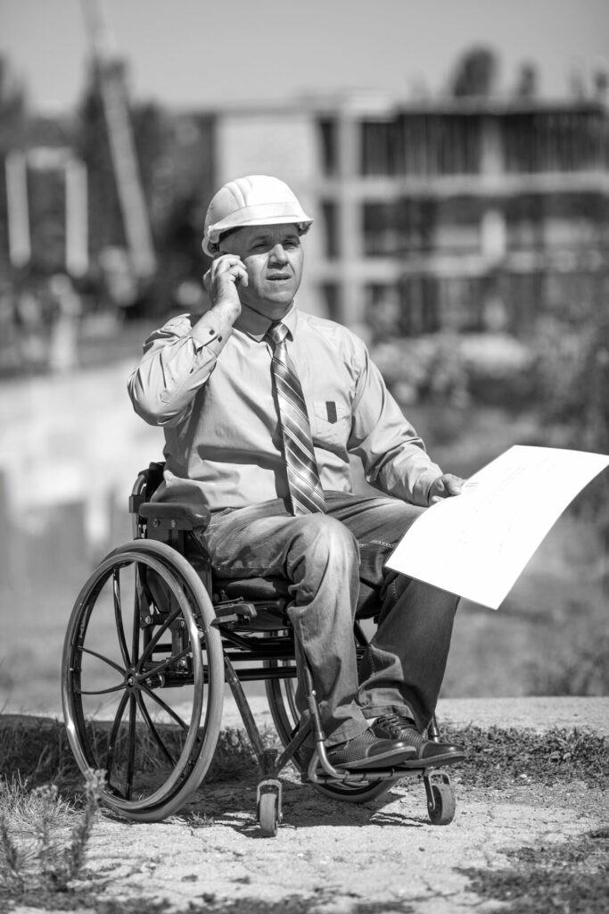 Disabled person in the construction helmet with documents in hand talking on the phone on the background of building. Successful wheelchair user directs the work.