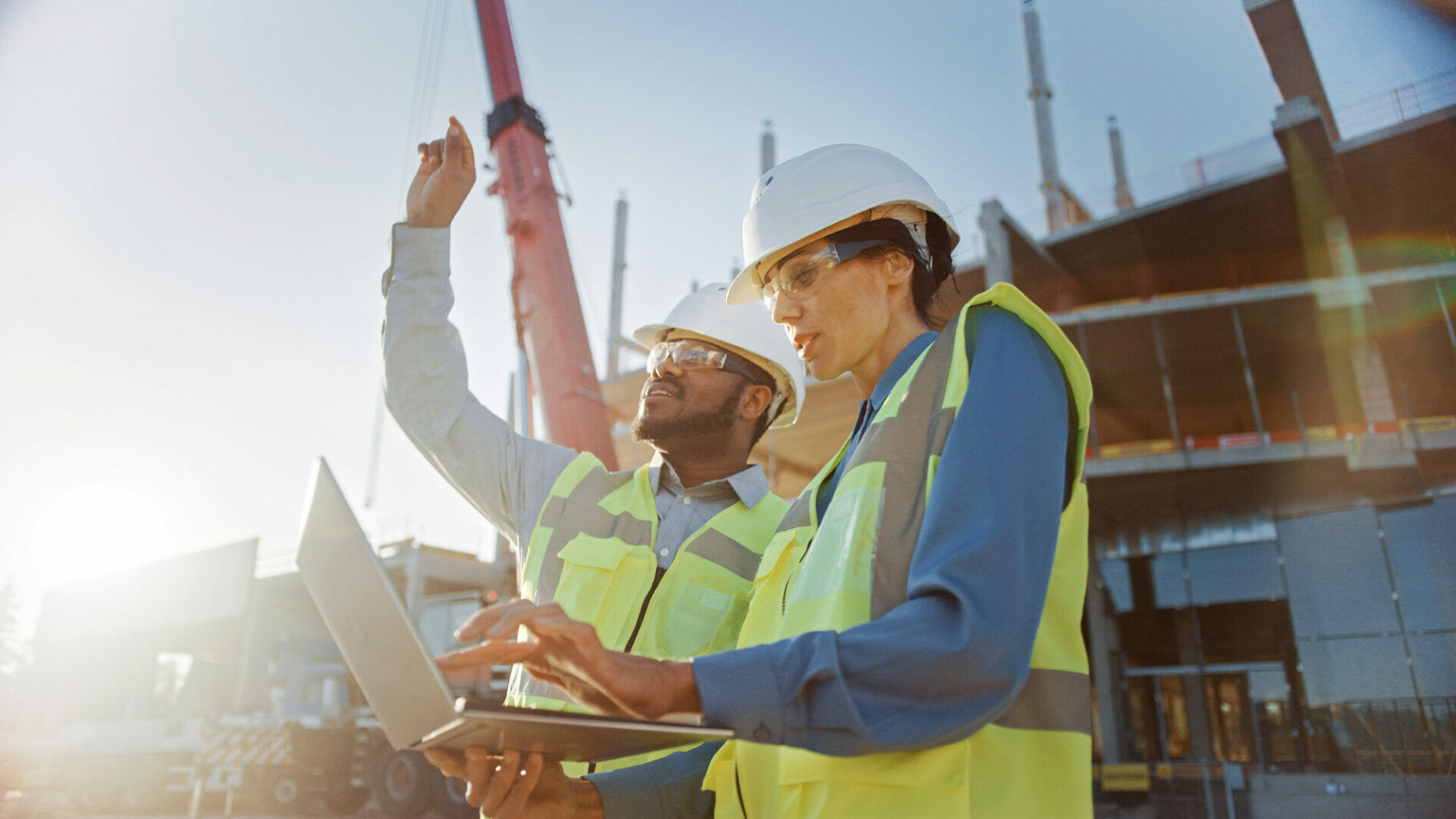 Two Specialists Inspect Commercial, Industrial Building Construction Site. Real Estate Project with Civil Engineer, Investor Use Laptop. In the Background Crane, Skyscraper Concrete Formwork Frames