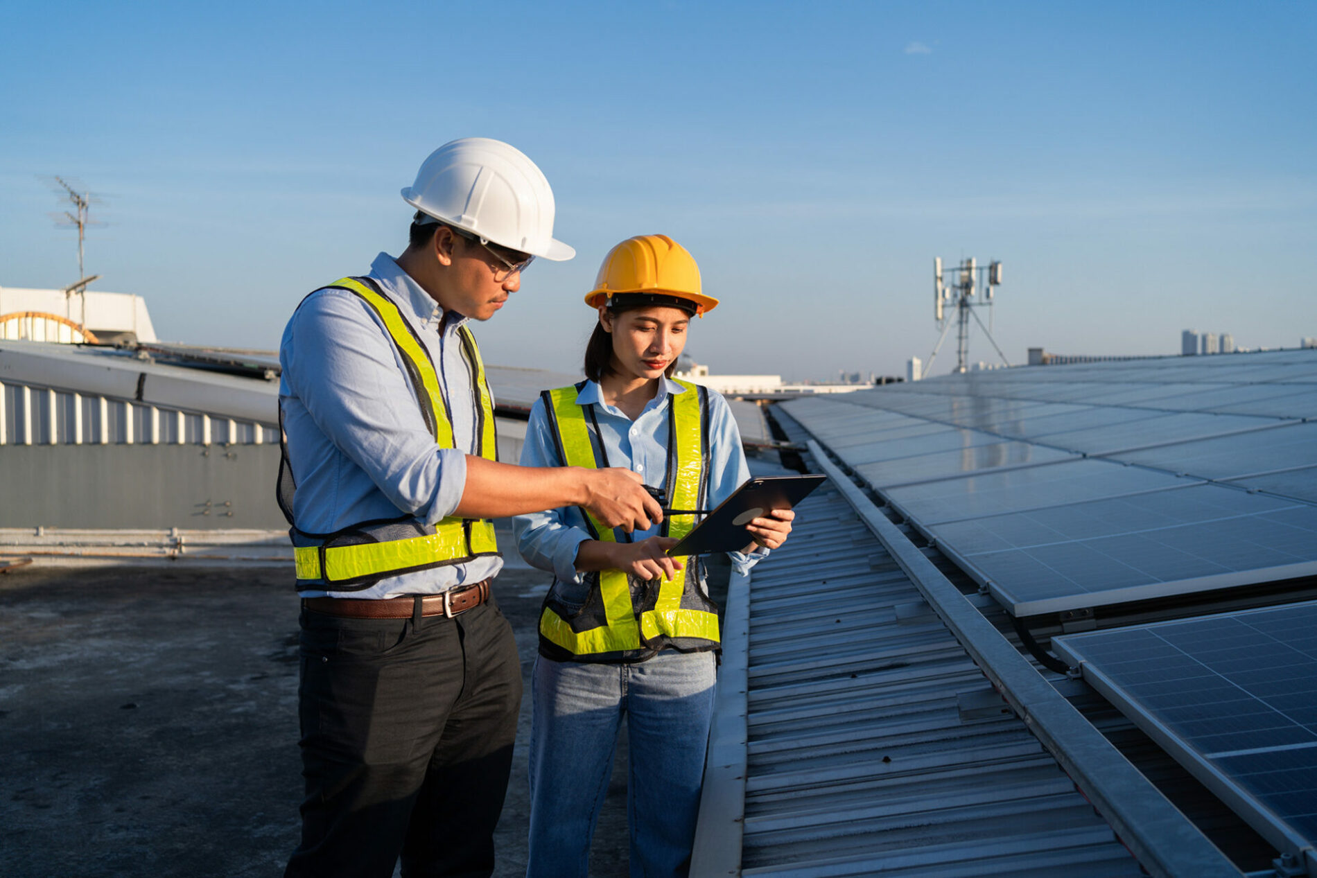 Two people are standing on a roof, one of them is pointing at a tablet. They are wearing safety vests and hard hats. Electrical engineer installs solar panels in power station