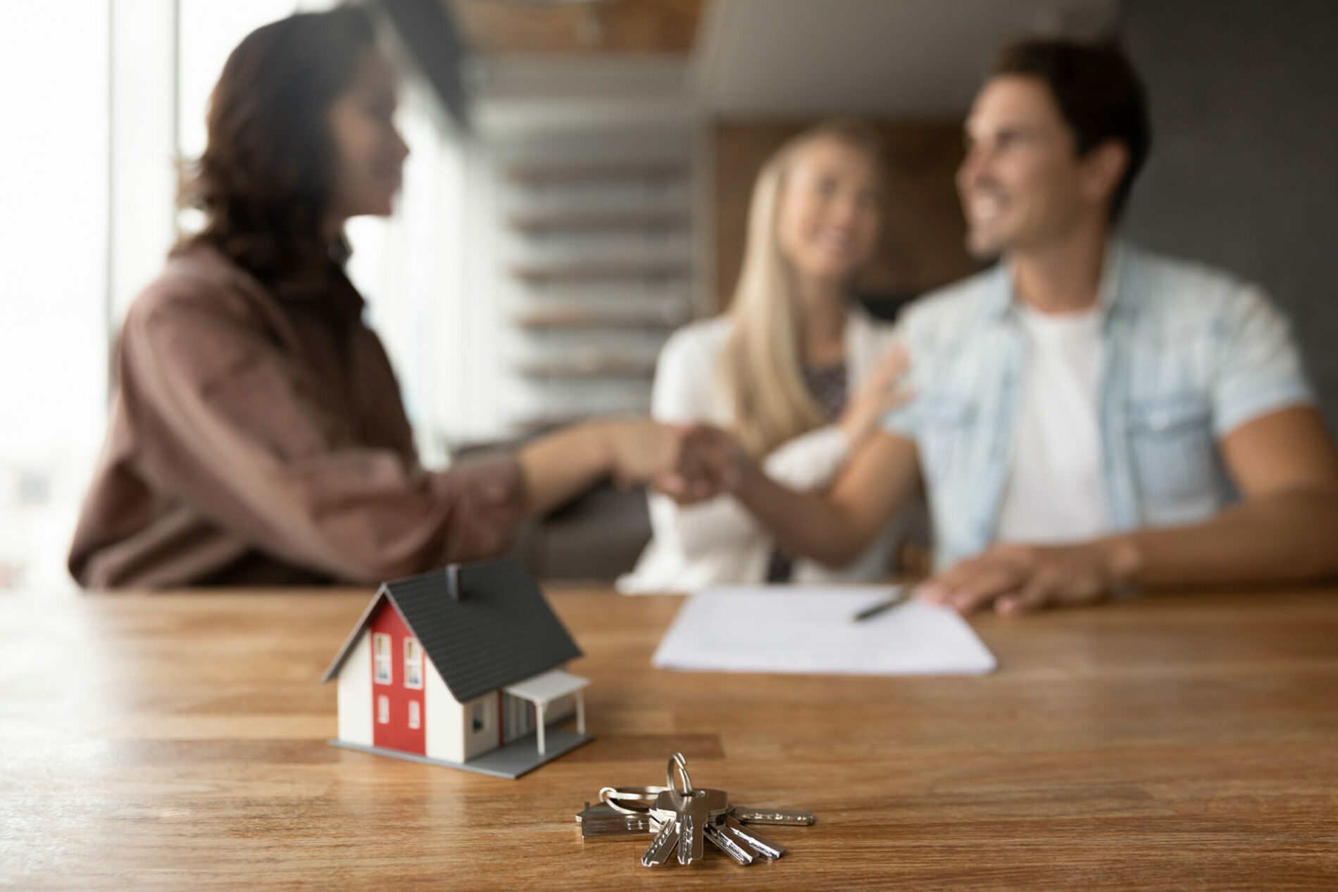 Close up of tiny toy house model and key on office table. Married couple of clients and real estate agent giving handshakes in office. Customers meeting with broker, seller for signing agreement